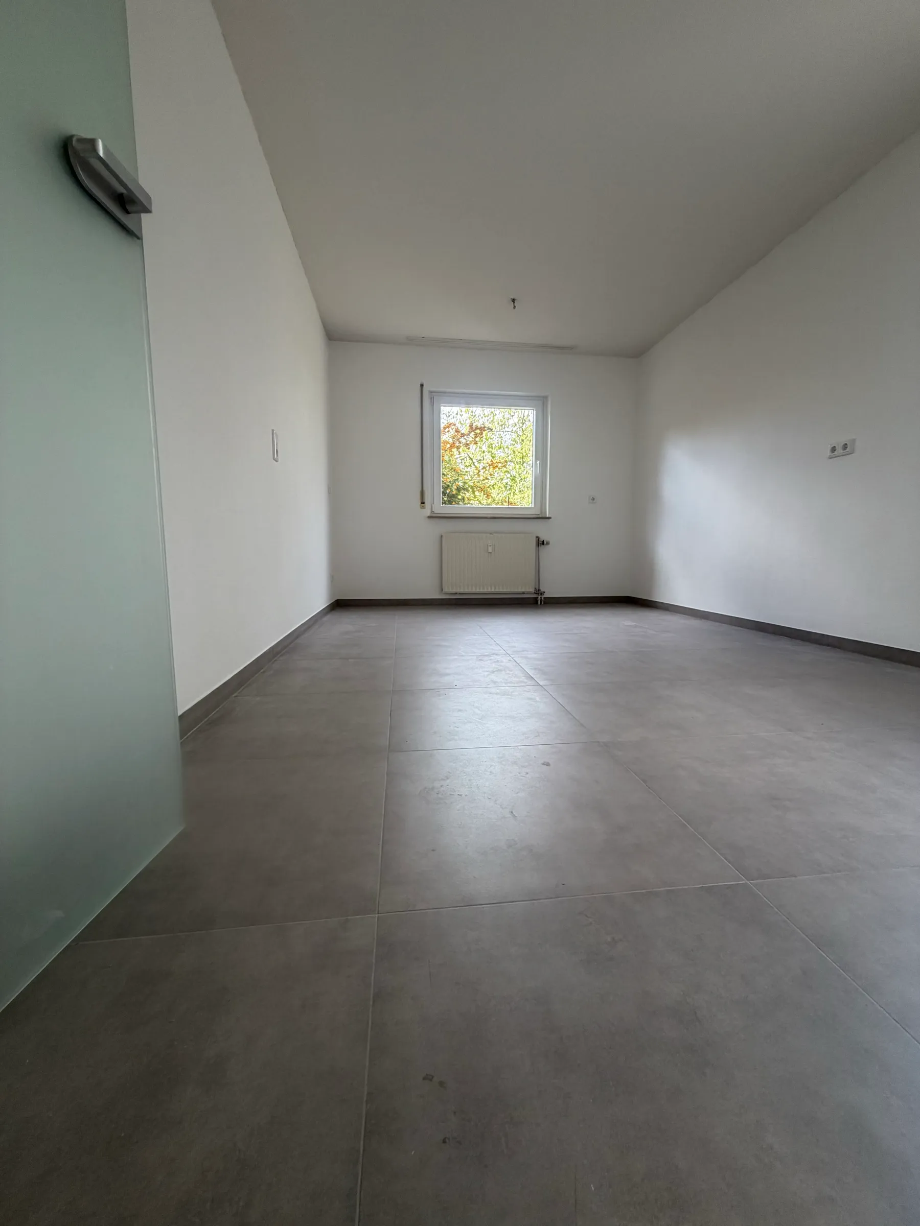 Dasselbe Zimmer nach der Sanierung: weiße Wände, graue Großformatfliesen, neue Glastür